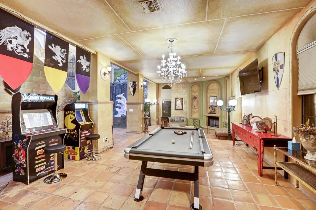 The game room at D&D Mansion in Vegas features high ceilings, a chandelier, and elegant antique furniture, with quirky touches like flags and a dolphin mural. A pool table dominates the center of the room, and arcade games Legends Ultimate and Pacman line the wall.
