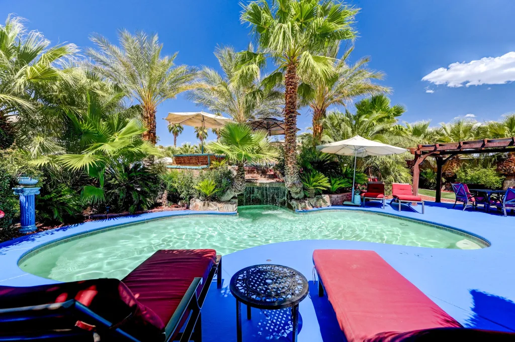 Two red lounge chairs offer a cozy spot for guests at Paradise Estate in Vegas to unwind in front of an aqua pool with tropical greens in the background,
