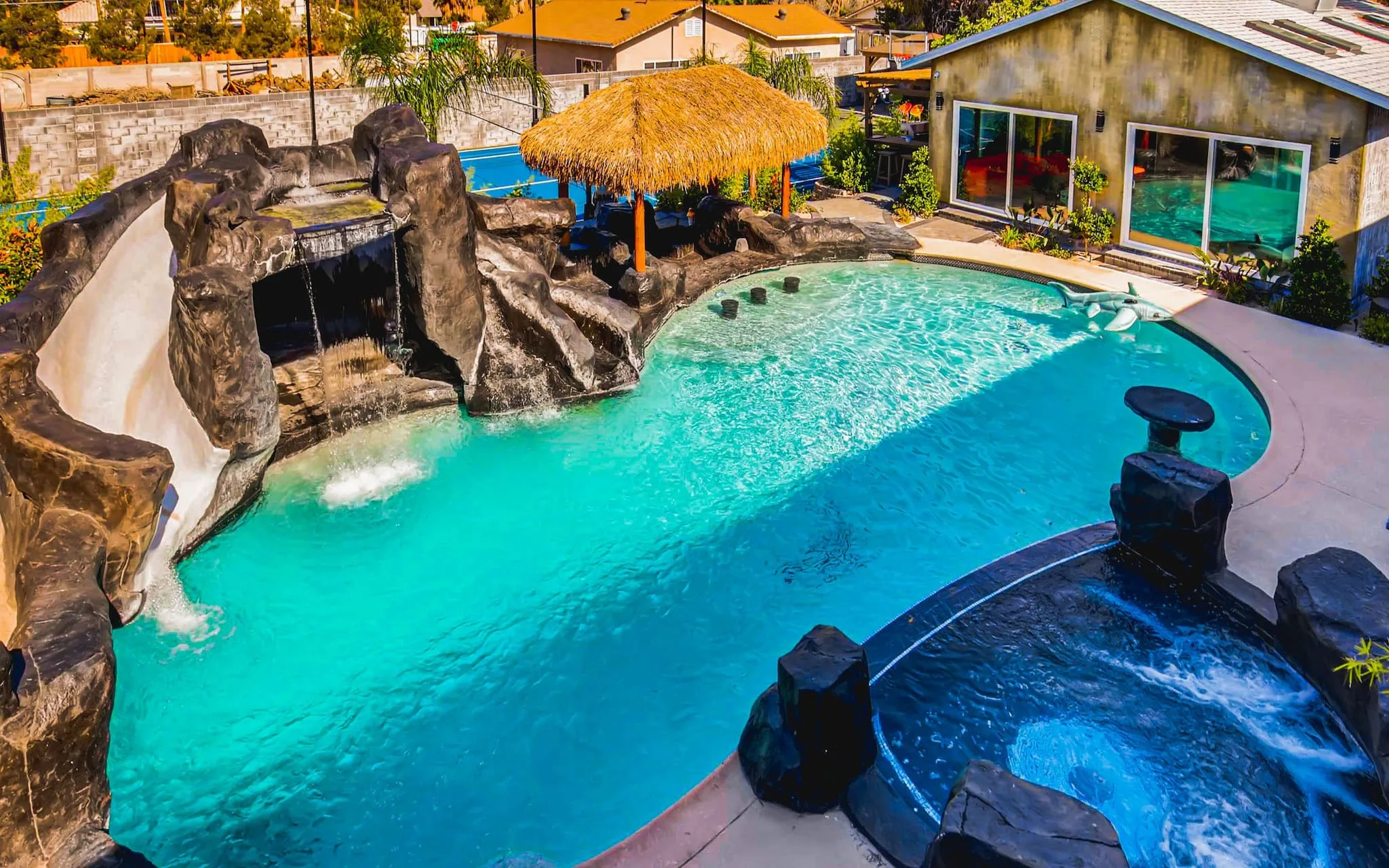 A luxury Vegas rental home is seen from above with a large pool at the centre, surrounded by tropical features like a groto, jacuzzi, palapas, slide and house.