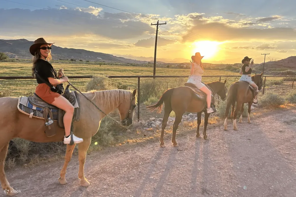 Three women sporting t-shirts, shorts and cowboy hats sit on horses while the sun sets behind them