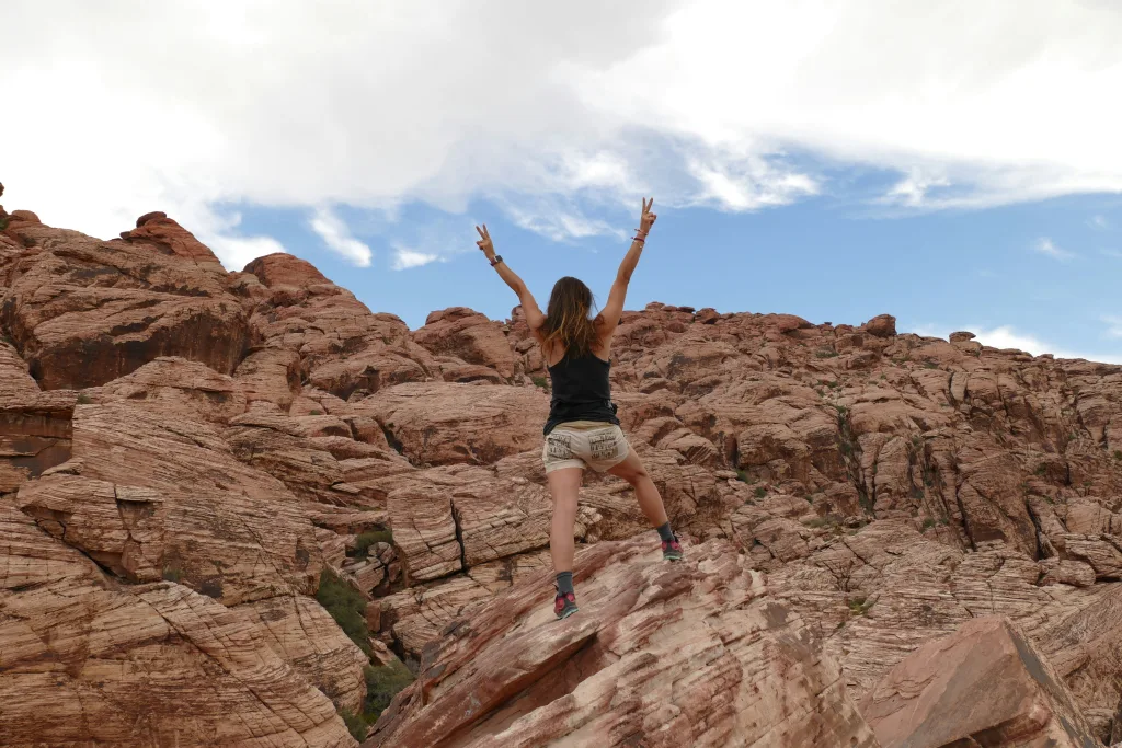 A woman wearing a black tank top, white shorts, and sneakers, stands on a rock formation facing away from the camera, arms in the air, making peace signs. The background is Red Canyon National Conservation Area, with a hill of reddish rocks and a blue sky with lots of white clouds.