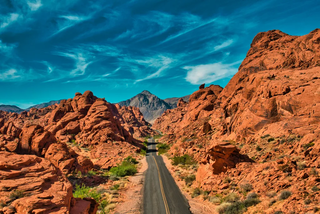 A black pavement road rolls up and down through a valley of red rock formations with a dramatic dark blue sky and mountains in the distance.