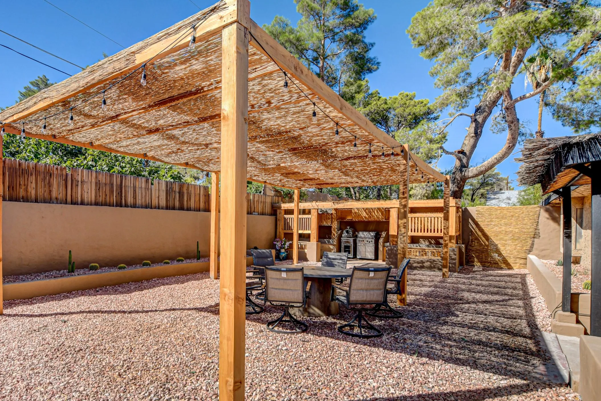 The backyard of Zen Haven, a luxury villa rental in Vegas, shows a wooden shade with a BBQ area and outdoor seating, and palm trees behind the fence.