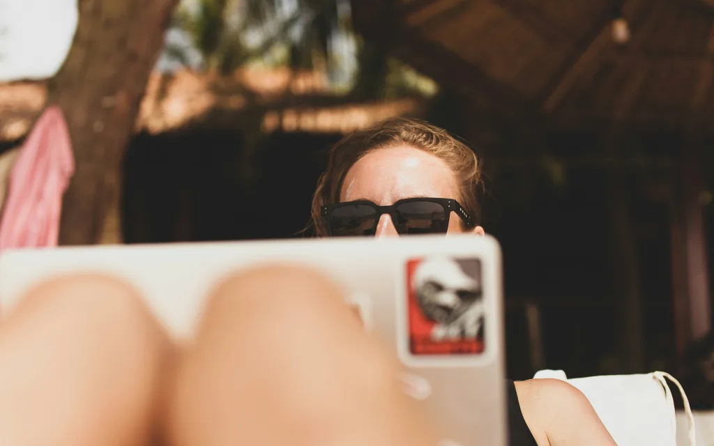 A woman looks at her laptop from a poolside lounger while wearing sunglasses