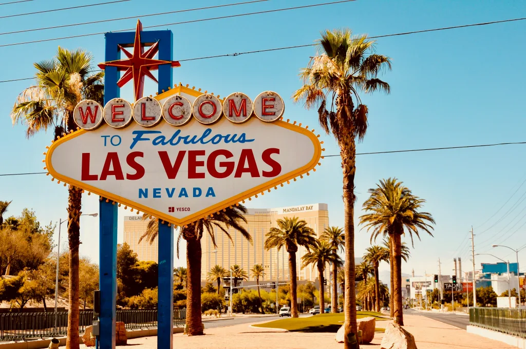 An iconic diamond-shaped vintage sign says "Welcome to Fabulous Las Vegas  Nevada" on the side of a Vegas road with a blue daytime sky. The  Mandalay can be seen in the background.
