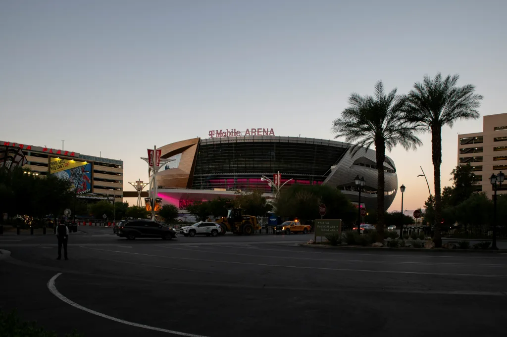 A round, flat-topped arena  is becoming a shadow as the sun goes down. The sign reads "T-Mobile Arena" nad some cars and palm trees can be seen on the Vegas road in front of it.
