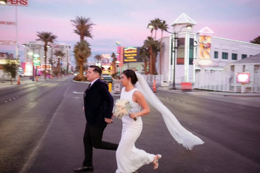 A man and woman couple in wedding attire hold hands as they cross a big road in Vegas, with palm trees in the background