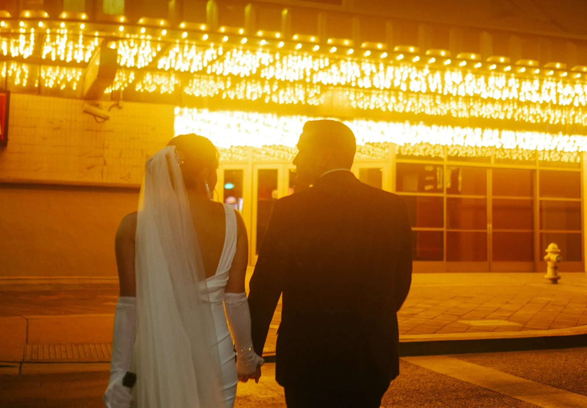 A man and woman in wedding attire look at each other while walking towards a building with soft red and white lighiting