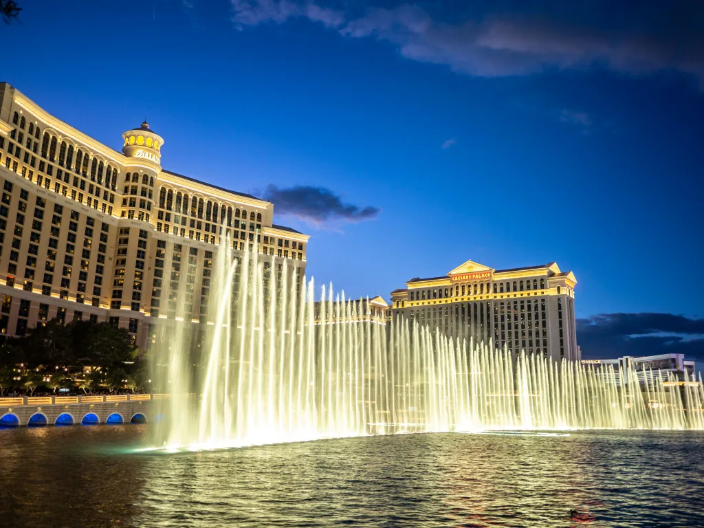 The Bellagio fountains reach for the sky in the foreground, with the hotel's classic style with arched windows behind to the left, and Caesars Palace roman-style hotel further back to the right.